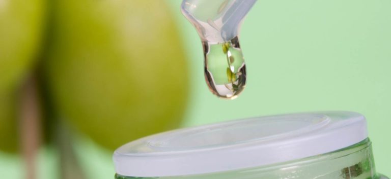 Olivenöl tropft in eine Flasche, im Hintergrund sind unscharf Oliven zu erkennen